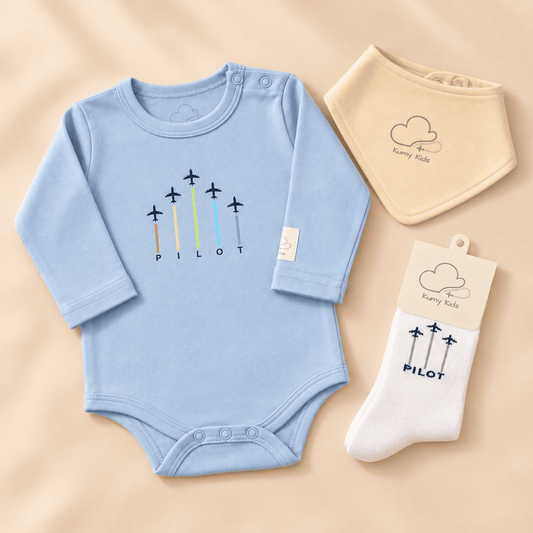 A baby clothing set laid flat on a warm beige background, including a light blue long-sleeve bodysuit with a colorful airplane and 'PILOT' graphic, a cream bib with a cloud and 'karry kids' logo, and a white sock in its paper packaging with the same cloud graphic and 'PILOT' text.
