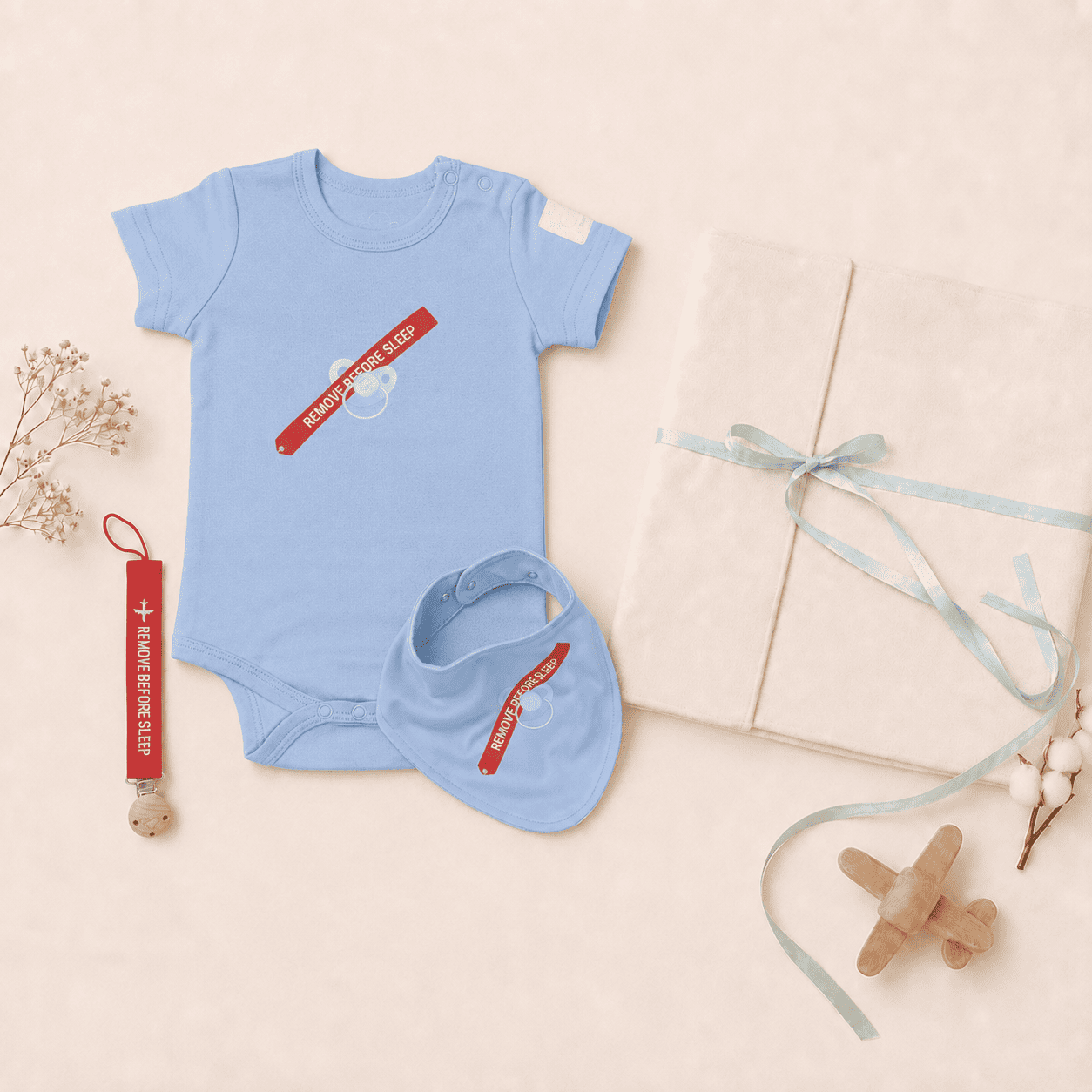 A baby bodysuit and matching bib set in light blue, styled in a flat lay with a red and white travel-style strap accessory, a cream gift box tied with a pale blue ribbon, dried flowers, and a small wooden airplane on a warm beige background.